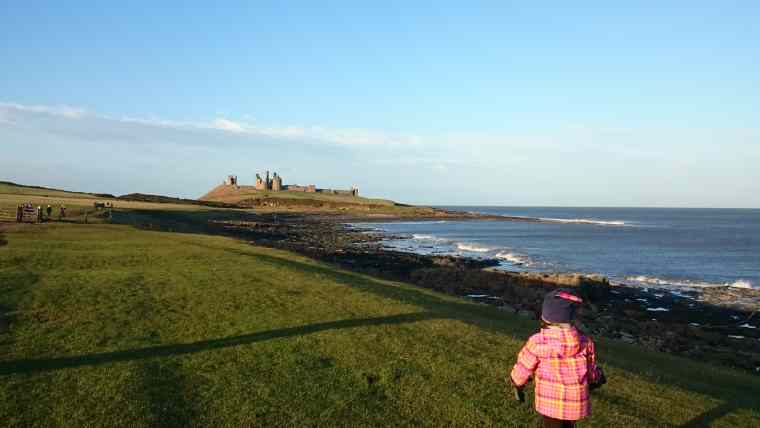 Image of girl in foreground and other people mid ground on grassy coastal path with castle and sea in background